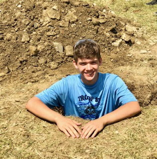 A young man, Noah McCauley, stands in a deep hole he has dug. The dirt is up to his chest. He is smiling.